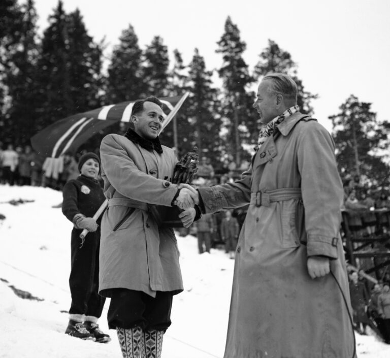 Hallgeir Brenden (t.v.) mottar Egebergs Ærespris på kulen etter 1. omgang av hopprennet i NM i 1954. Det er Arthur Ruud som overrakte prisen. I bakgrunnen en lokal flaggbærer.