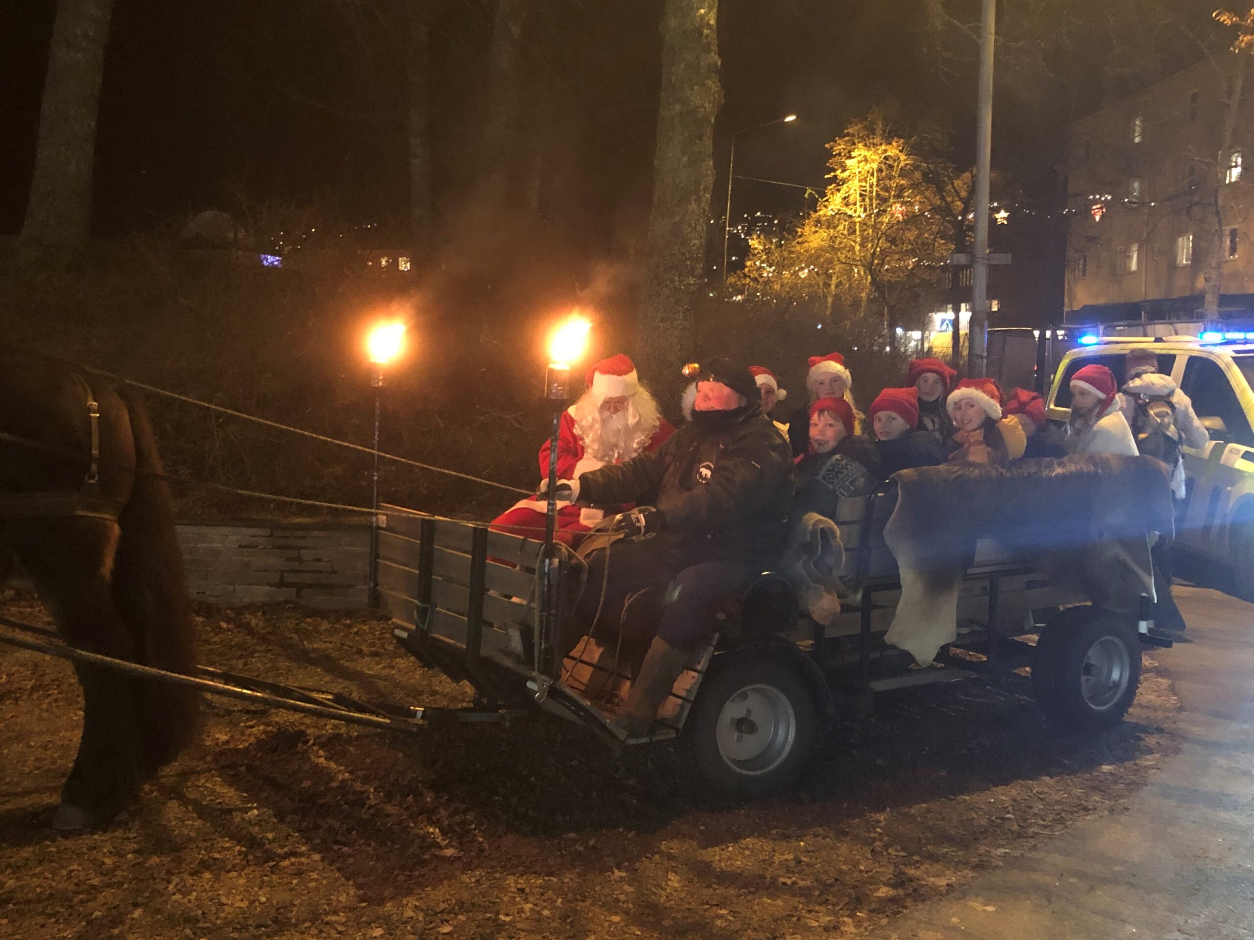 Julenissen har med seg barna på sleden sin. I bakgrunnen er lysene tent i julegata.