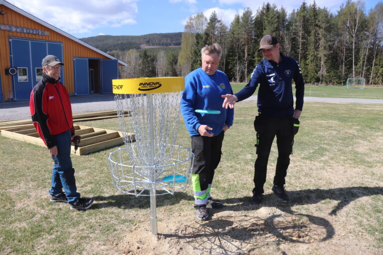 Tom Solås (f.v.), leder av sommeranleggskomiteen i Brandval IL, Ove Jonny Løvberget og Sondre Skulstad demonstrerer frisbeegolfkurv på Idrettsplassen ved Brandval. Den nye banen har ni slike.