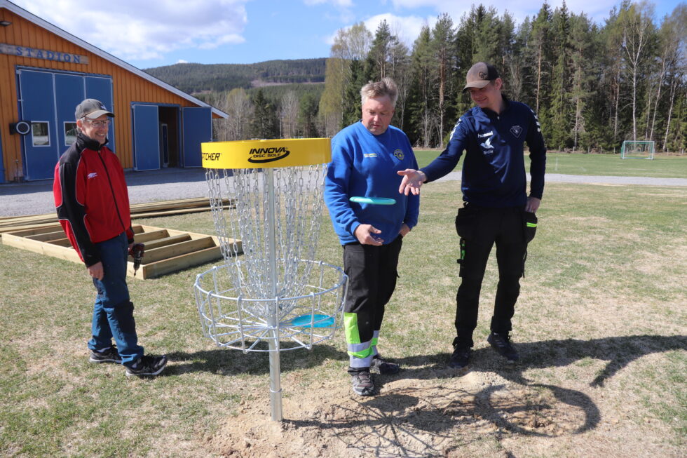 Tom Solås (f.v.), leder av sommeranleggskomiteen i Brandval IL, Ove Jonny Løvberget og Sondre Skulstad demonstrerer frisbeegolfkurv på Idrettsplassen ved Brandval. Den nye banen har ni slike.