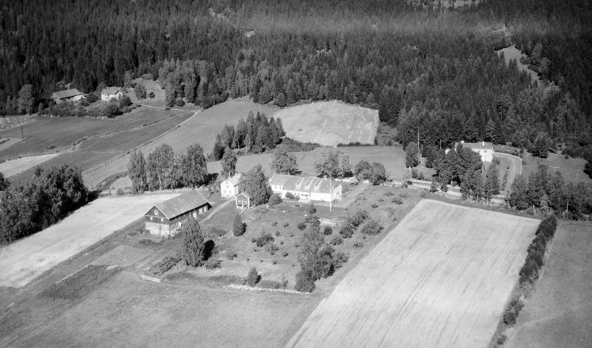 Galgebakken lå under gården Langeland Øvre, som hadde grense mot Søndre Tråstad omtrent midt i Øvrebyen for kjøpstaden ble skilt ut i 1974. Bilde tatt av Vilhelm Skappel i 1950 for Widerøe. Langeland skole ligger til høyre for hovedbygningen i dag. Foto: Nasjonalbiblioteket