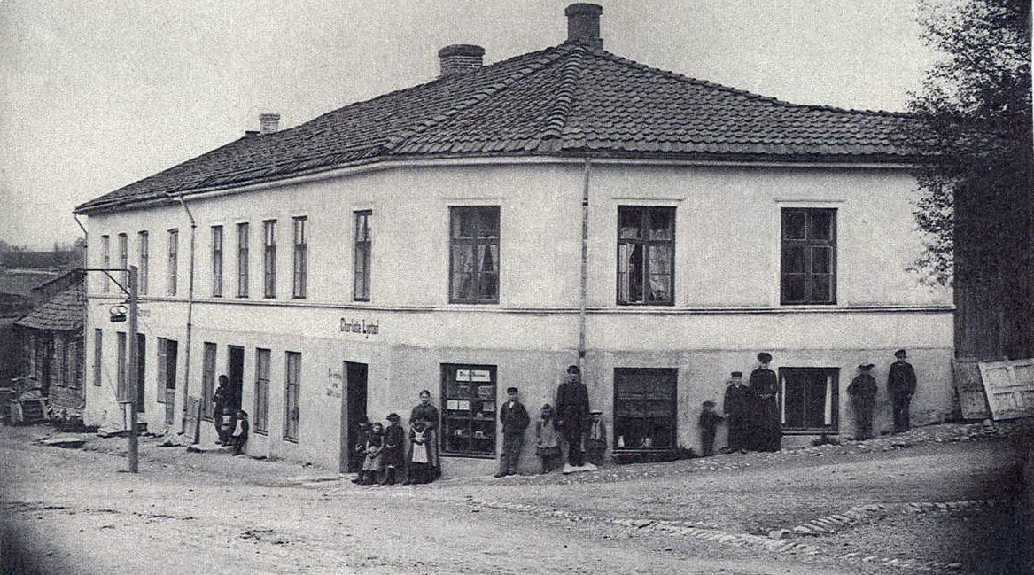Holmengården ble bygget av Halvor Olsen i 1858. Her bodde familien Schmidt ved folketellingen i 1865 og her lå også salongen til Johan Fredrik. Charlotte Lystad fra Elverum tilbød bevertning med kaffe og mat på 1880-tallet, som skiltet viser. Den staslige murgården brant i 1885 og ble bygget opp i tre etasjer.
