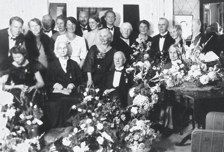 Familien Gulliksen/Schulerud, her trolig samlet ved Sigrids svoger Gunvald Gulliksen (sittende foran) sin 80-årsdag. Sigrid står bak ham til høyre, med kort mørkt hår, bak den høyeste blomsteroppsatsen.